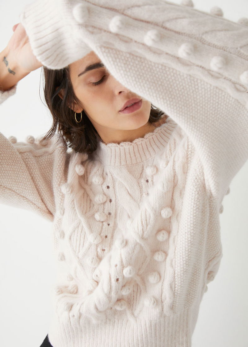 Cream cable-knit sweater with oversized, textured sleeves and decorative pom-poms.