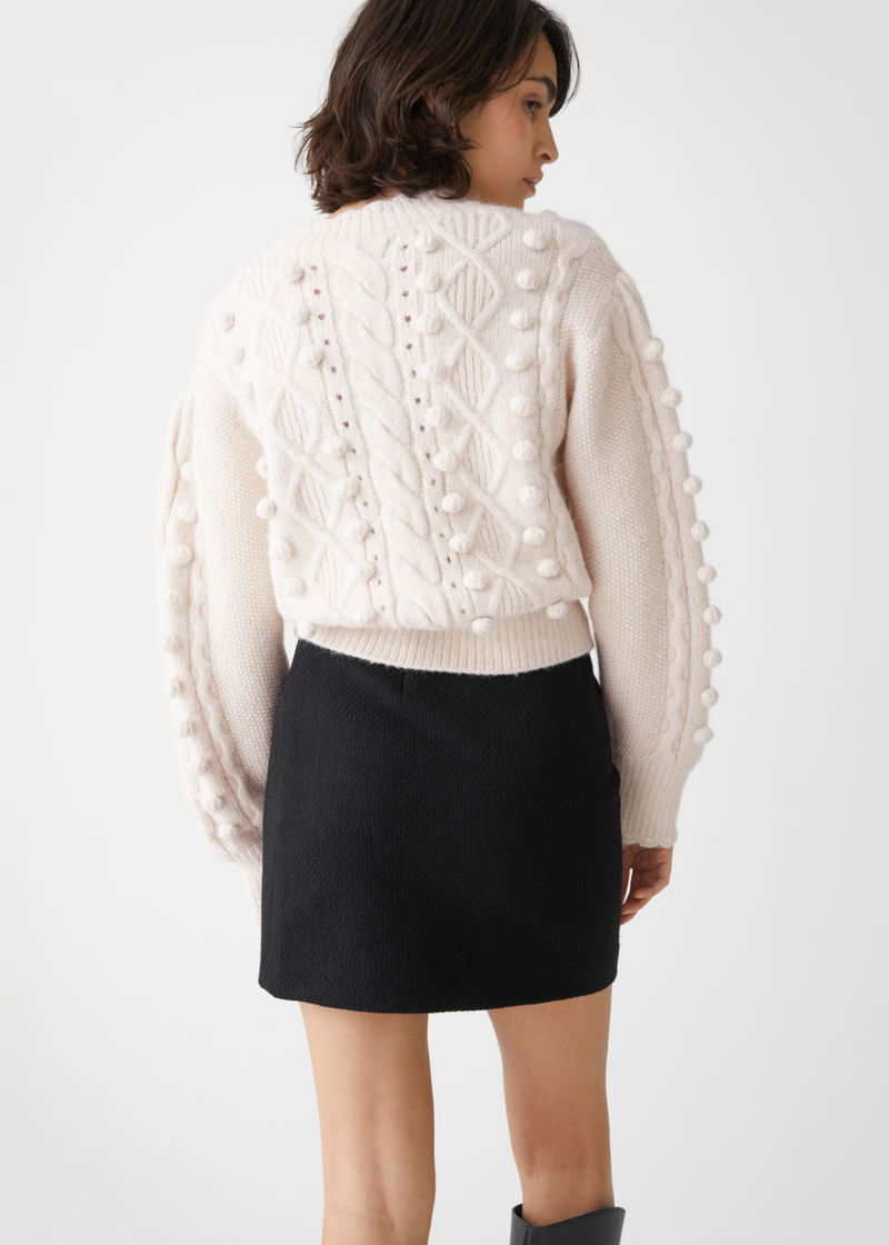 Cream cable-knit sweater with pom-poms, worn with a black pleated mini skirt and black boots.