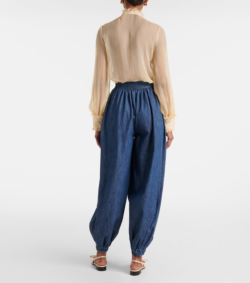 Valentino: Beige sheer blouse with ruffled sleeves paired with wide-leg, drawstring denim pants.