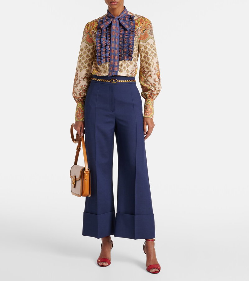 Valentino model wearing a paisley print blouse with ruffled neckline and chain belt, paired with wide-leg navy pants and heels.