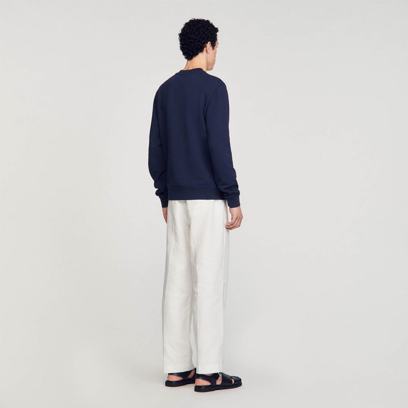 Man wearing a navy sweatshirt and white trousers, sandals.