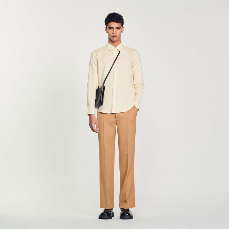 Man wearing a beige shirt, brown trousers, and black shoes, holding a black crossbody bag.