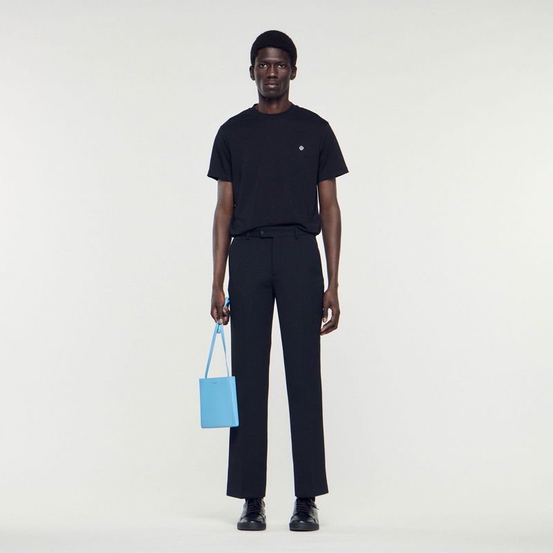 Man wearing a black t-shirt and tailored trousers, holding a light blue bag.