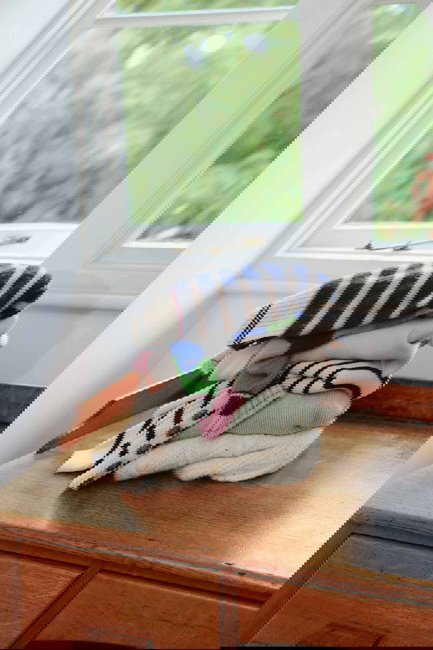 A pile of colourful, folded jumpers on a wooden desk by a window