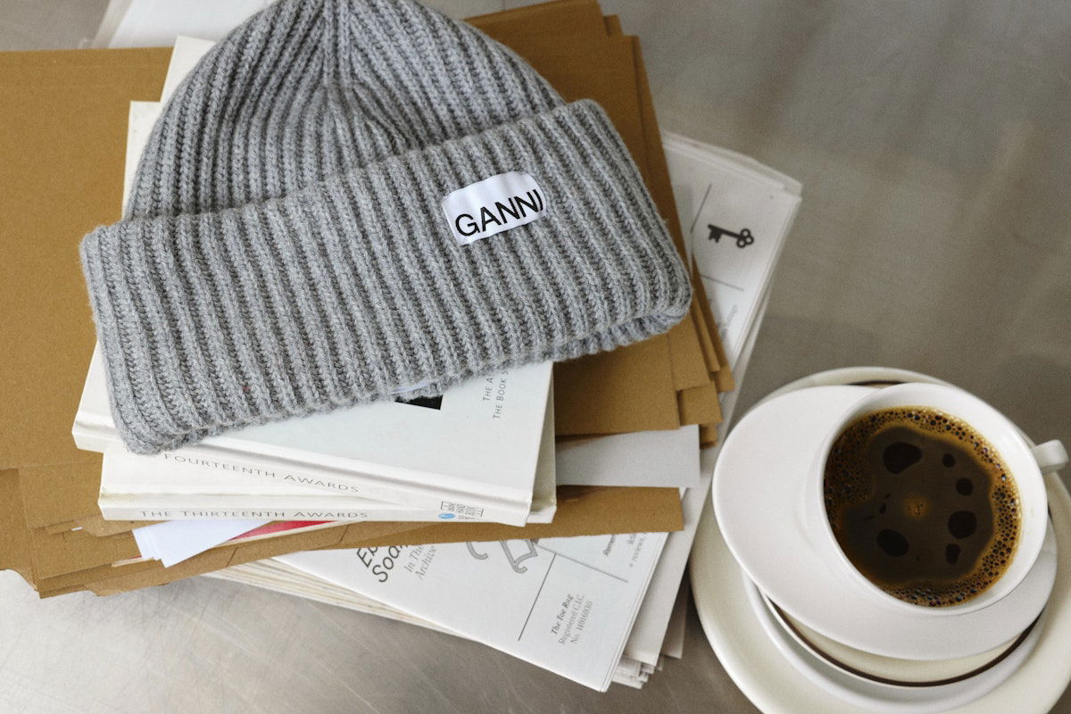 Grey Ganni beanie on a pile of paper and envelopes, next to a white cup of black coffee